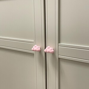 Op de afbeelding: Twee lichtroze wolkvormige kastknoppen op een lichtgrijze kastdeur. De knoppen zijn gecentreerd en geven een speelse uitstraling. De kast is lichtgrijs geschilderd.