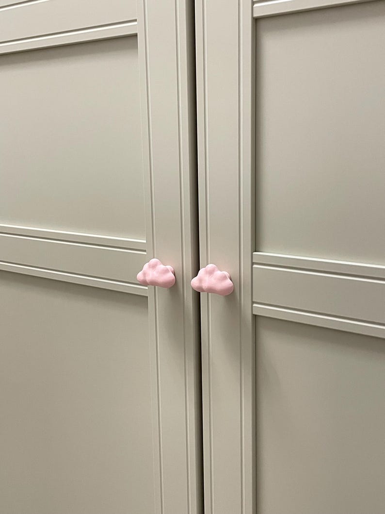 Op de afbeelding: Twee lichtroze wolkvormige kastknoppen op een lichtgrijze kastdeur. De knoppen zijn gecentreerd en geven een speelse uitstraling. De kast is lichtgrijs geschilderd.