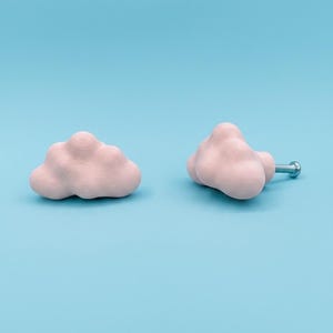 Op de afbeelding: Twee roze wolkvormige kastknoppen op een lichtblauwe achtergrond. E&eacute;n knop is een losse wolk, de andere heeft een zilveren schroef. De knoppen zijn gemaakt van een textuurrijk materiaal, wat een unieke touch geeft aan elk meubel.