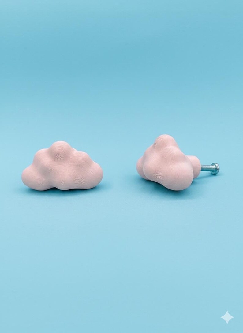 Op de afbeelding: Twee roze wolkvormige kastknoppen op een lichtblauwe achtergrond. E&eacute;n knop is een losse wolk, de andere heeft een zilveren schroef. De knoppen zijn gemaakt van een textuurrijk materiaal, wat een unieke touch geeft aan elk meubel.