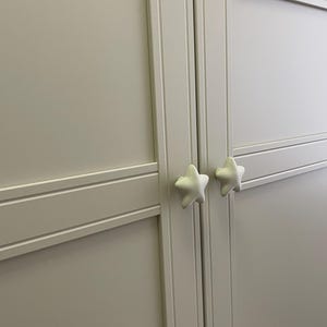 May include: A light grey cabinet door with two white star-shaped knobs. The door features recessed panels and horizontal and vertical trim. The knobs are positioned centrally on the door.
