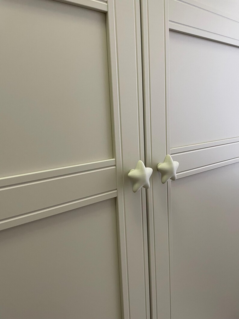 May include: A light grey cabinet door with two white star-shaped knobs. The door features recessed panels and horizontal and vertical trim. The knobs are positioned centrally on the door.