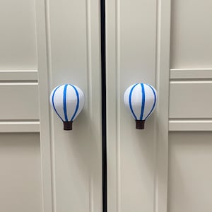 May include: Two white cabinet knobs shaped like hot air balloons, each with blue stripes and a brown base. The knobs are mounted on a light-colored cabinet door. The knobs are decorative and functional.
