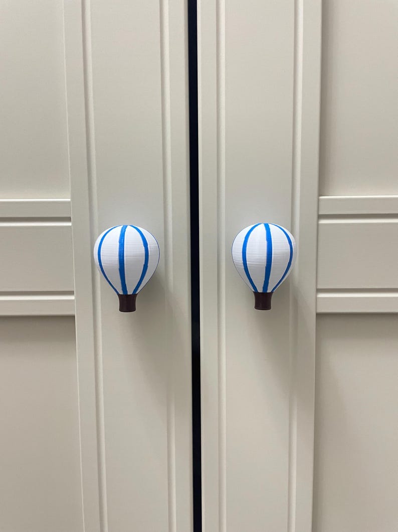 May include: Two white cabinet knobs shaped like hot air balloons, each with blue stripes and a brown base. The knobs are mounted on a light-colored cabinet door. The knobs are decorative and functional.
