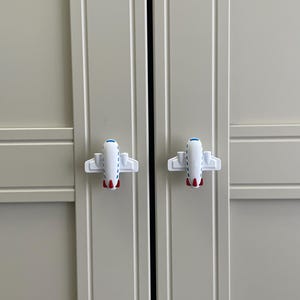 May include: Two white airplane-shaped cabinet knobs with blue and red accents are attached to a light gray cabinet door. The knobs are symmetrical and add a playful touch to the cabinet.