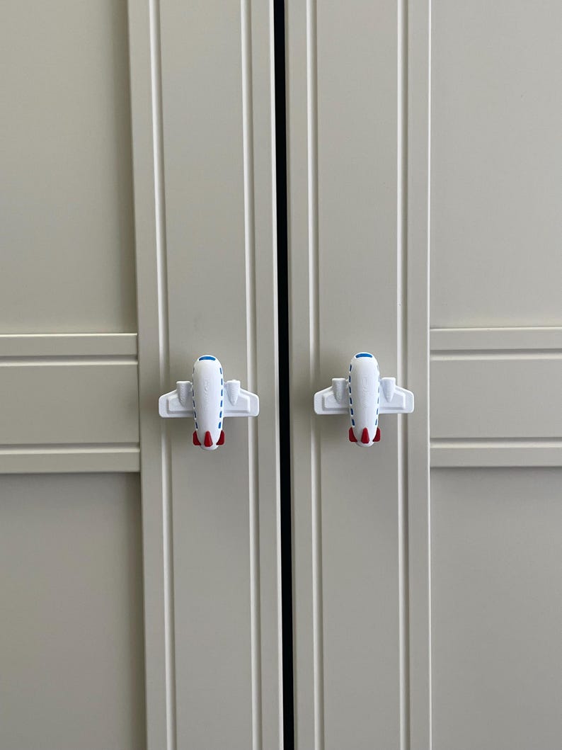 May include: Two white airplane-shaped cabinet knobs with blue and red accents are attached to a light gray cabinet door. The knobs are symmetrical and add a playful touch to the cabinet.
