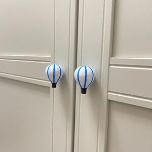 May include: Two white cabinet knobs shaped like hot air balloons with blue stripes and brown bases. The knobs are mounted on a light-colored cabinet door. The knobs add a whimsical touch to furniture.