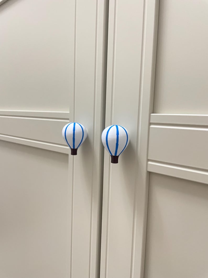 May include: Two white cabinet knobs shaped like hot air balloons with blue stripes and brown bases. The knobs are mounted on a light-colored cabinet door. The knobs add a whimsical touch to furniture.