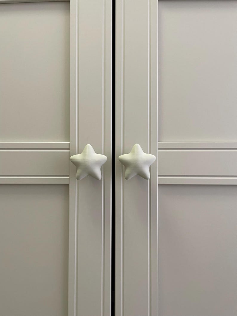 May include: A light grey cabinet with two doors, each featuring a white, star-shaped knob. The knobs have a textured surface. The cabinet doors have vertical panels. The design is simple and functional, suitable for storage.