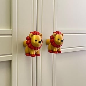 Puede incluir: Dos tiradores de armario con forma de leones de dibujos animados. Los leones son amarillos con melenas, patas y detalles rojos. Los tiradores están montados en una puerta de armario blanca. Los tiradores son decorativos y funcionales.