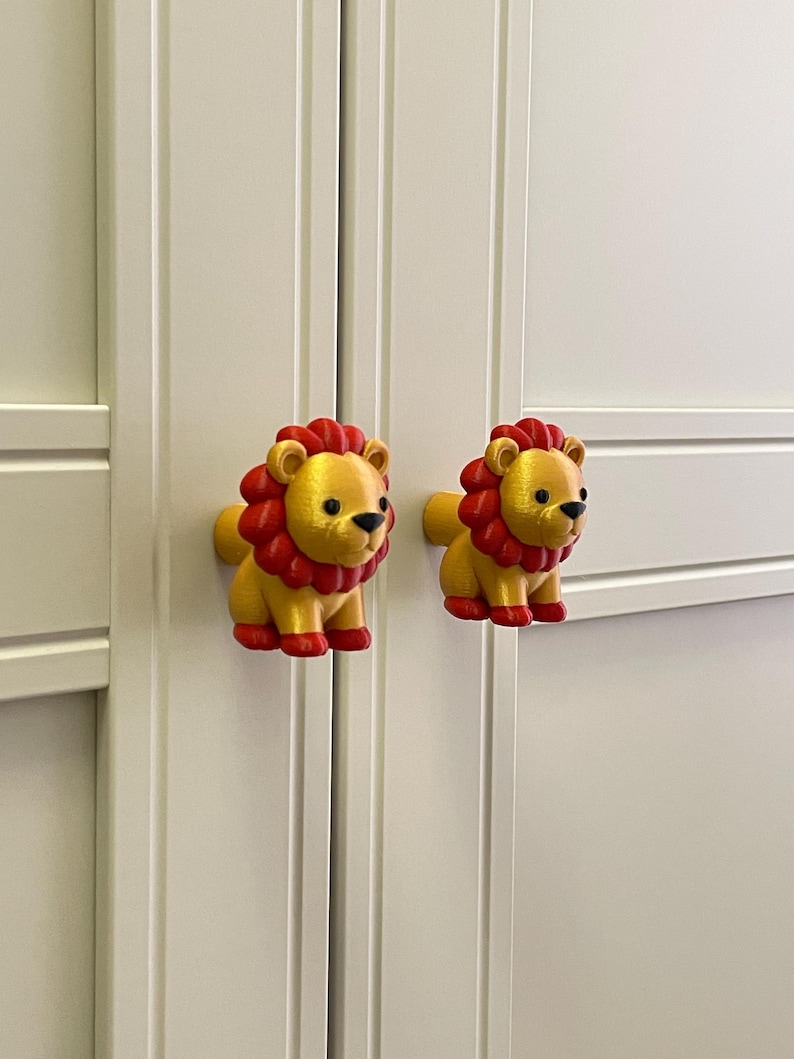 Pu&ograve; includere: Due pomelli per armadi a forma di leoni dei cartoni animati. I leoni sono gialli con criniere, zampe e dettagli rossi. I pomelli sono montati su un'anta bianca. I pomelli sono decorativi e funzionali.