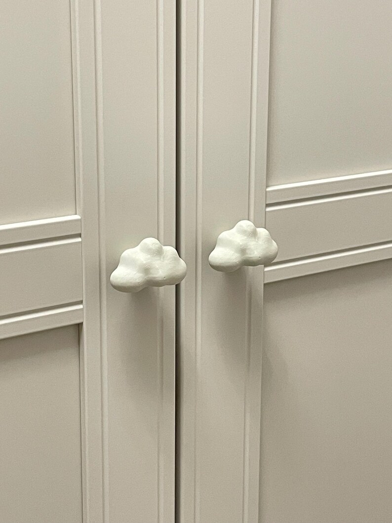 Op de afbeelding: Een close-up van een witte kast met twee wolkvormige handgrepen. De kastdeuren zijn lichtbeige van kleur. De wolkenknoppen zijn wit en getextureerd, wat een speelse toets aan het meubel geeft.
