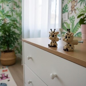 Peut inclure: Deux boutons de meuble en forme de girafe avec des taches noires et de petites cornes sont pos&eacute;s sur une surface en bois. Les boutons sont fix&eacute;s &agrave; une commode blanche &agrave; trois tiroirs. La pi&egrave;ce a un papier peint sur le th&egrave;me de la jungle.
