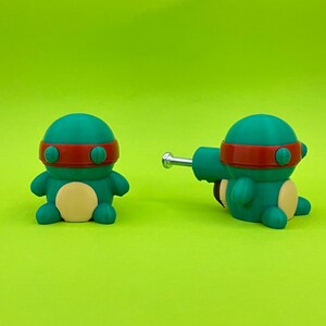 K&ouml;nnte beinhalten: Zwei teal- und rotfarbene Cartoon-Schildkr&ouml;tenfiguren mit hellgelbem Bauch und rotem Stirnband. Eine Figur steht aufrecht, die andere liegt mit einer silbernen Metallstange an der Seite. Der Hintergrund ist einfarbig gr&uuml;n.