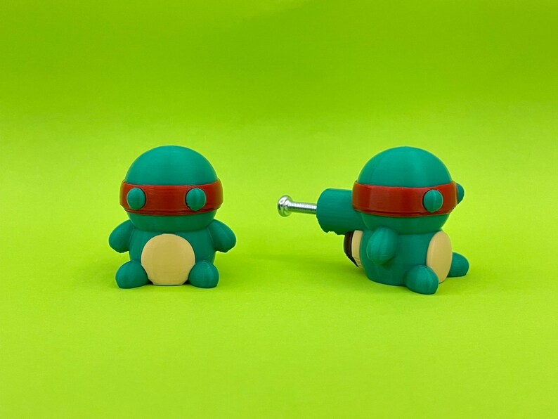 K&ouml;nnte beinhalten: Zwei teal- und rotfarbene Cartoon-Schildkr&ouml;tenfiguren mit hellgelbem Bauch und rotem Stirnband. Eine Figur steht aufrecht, die andere liegt mit einer silbernen Metallstange an der Seite. Der Hintergrund ist einfarbig gr&uuml;n.