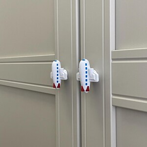 May include: Two white airplane-shaped cabinet knobs with blue and red accents are attached to a light gray cabinet. The knobs are designed to look like small airplanes, adding a playful touch to the furniture.