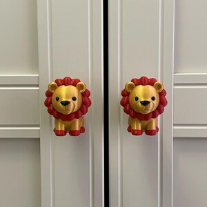 Pu&ograve; includere: Due pomelli per armadi a forma di leone dorato con criniere e zampe rosse sono attaccati a una porta di un armadio bianco. Le manopole sono simmetriche e aggiungono un tocco giocoso.