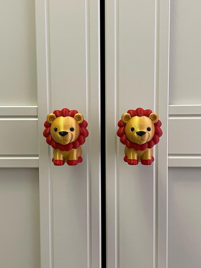 Pu&ograve; includere: Due pomelli per armadi a forma di leone dorato con criniere e zampe rosse sono attaccati a una porta di un armadio bianco. Le manopole sono simmetriche e aggiungono un tocco giocoso.