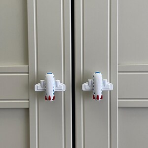 Könnte beinhalten: Zwei weiße, flugzeugförmige Schrankknöpfe mit roten und blauen Akzenten sind an einer hellgrauen Schranktür befestigt. Die Knöpfe sind symmetrisch und verleihen dem Schrank eine verspielte Note.