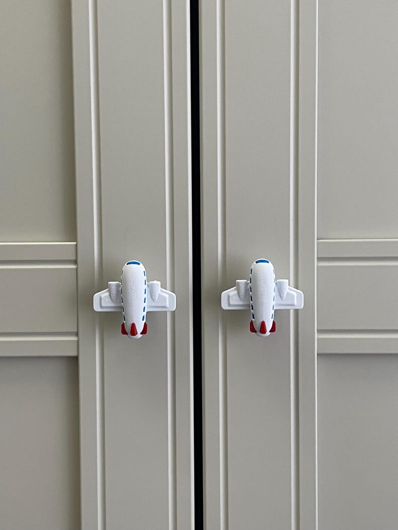 May include: Two white airplane-shaped cabinet knobs with red and blue accents are attached to a light gray cabinet door. The knobs are symmetrical and add a playful touch to the cabinet.
