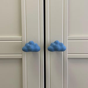 May include: Two light blue cloud-shaped cabinet knobs on a white cabinet door. The knobs have a soft, rounded shape, adding a whimsical touch. The cabinet doors have vertical paneling. The knobs are likely made of ceramic.