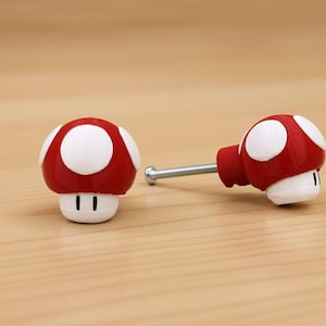 以下が含まれることがあります： 赤と白のキノコ型のキャビネットノブが2つ。1つは直立しており、もう1つは横になっています。ノブは白いベースと赤いキャップに白い斑点があります。銀色のネジがキノコの側面から伸びています。