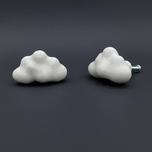 Op de afbeelding: Twee witte wolkvormige kastknoppen. E&eacute;n knop ligt plat, terwijl de andere rechtop staat, met een zilveren schroef bevestigd. De knoppen hebben een getextureerd oppervlak en staan tegen een donkergrijze achtergrond.