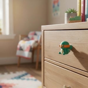 K&ouml;nnte beinhalten: Ein gr&uuml;ner und orangefarbener Cartoon-Schildkr&ouml;ten-Knauf an einer hellen Holzkommode. Die Kommode befindet sich in einem Kinderzimmer, mit einer bunten Steppdecke und Spielzeug im Hintergrund.