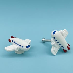 May include: Two white airplane-shaped cabinet knobs with blue and red accents. One knob is mounted on a silver screw. The knobs have a whimsical design, perfect for a child's room or a travel-themed space.