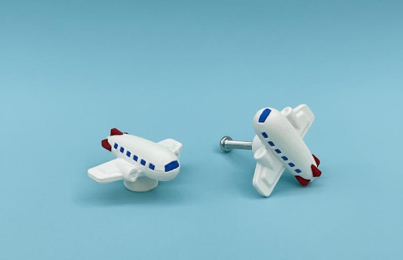 May include: Two white airplane-shaped cabinet knobs with blue and red accents. One knob is mounted on a silver screw. The knobs have a whimsical design, perfect for a child's room or a travel-themed space.