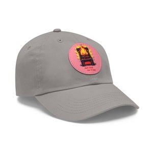 Può includere: Un cappellino da baseball grigio con visiera curva. Il cappellino presenta una toppa circolare con un'immagine a cartoni animati di un cabinato arcade in fiamme su sfondo rosa. Il testo "ARCADE ON FIRE" è stampato sotto l'immagine.