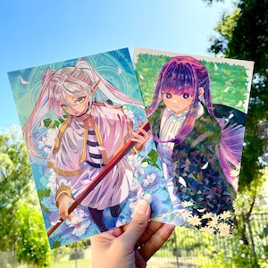 May include: Two anime-style art prints held up against a bright blue sky. One print features a character with white hair and green eyes, holding a staff. The other print shows a character with purple hair. Both prints have floral backgrounds.