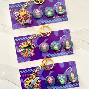 May include: Three sets of acrylic keychains and pins featuring Splatoon-inspired weapon designs on yellow ink splatters, accompanied by three circular pins depicting pink, green, and orange squid characters on purple display cards with geometric patterns.