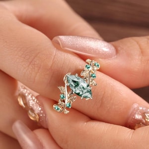 May include: A gold ring featuring a central marquise-cut moss agate stone, flanked by intricate metal leaf motifs accented with small, round, vibrant green gemstones set along the band, creating a botanical-inspired design on the jewelry piece.