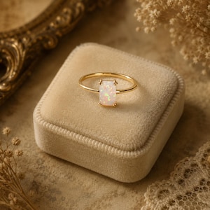 May include: A gold ring featuring a rectangular, iridescent white opal stone set in a four-prong setting. The band is thin and polished, showcasing a smooth, metallic finish that complements the light-reflecting properties of the central gemstone.