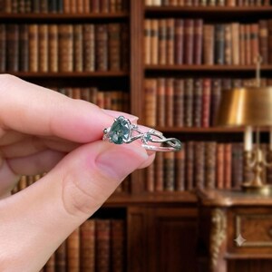 May include: A silver-toned ring featuring a pear-shaped moss agate gemstone as the central focus. The band is designed with a delicate, intertwining vine-like structure that incorporates small, clear accent stones along the metal branches.