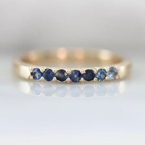 May include: A gold band ring featuring a series of seven round, dark blue gemstones. The ring has a smooth, polished finish and is set against a soft, blurred background. The gemstones are evenly spaced along the band.
