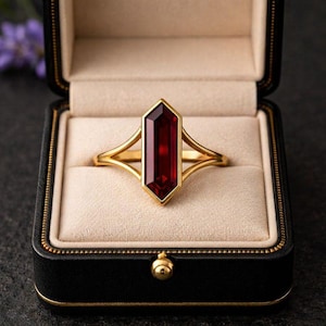 May include: A gold ring with a rectangular, faceted, deep red gemstone. The ring is set in a gold band with a unique open design. The ring is displayed in a black jewelry box with a gold clasp and a cream-colored interior.