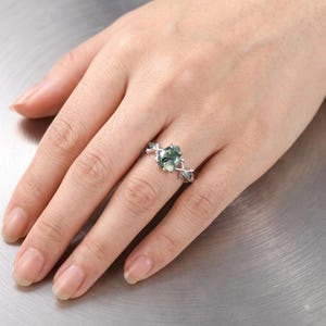 May include: A silver-toned ring featuring a pear-shaped moss agate center stone with a green and translucent appearance. The band is designed with a branch-like motif, incorporating small, round, green accent stones on either side of the center.