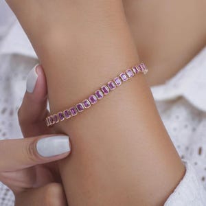 Puede incluir: Una pulsera de oro rosa con una fila de piedras preciosas rosas de corte cuadrado. La pulsera se lleva en la muñeca, con las piedras preciosas muy juntas. El fondo es una tela blanca texturizada.