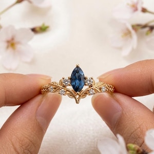 May include: A gold ring featuring a central marquise-cut dark blue gemstone flanked by four smaller round clear stones. The band is designed with intricate leaf motifs and a textured finish, creating a botanical aesthetic across the metal surface.