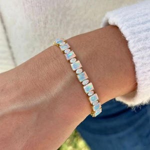 May include: A gold bracelet with rectangular and oval opals. The opals display a milky white base with blue and green flashes. The bracelet is worn on a wrist, highlighting the jewellery's design and colour.
