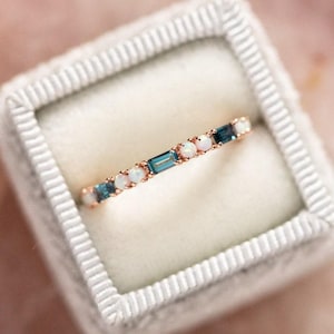 Op de afbeelding: Een roségouden ring met afwisselend blauwe en witte edelstenen. De ring heeft rechthoekige en ronde stenen in een delicate band. De ring wordt gepresenteerd in een witte fluwelen ringdoos.