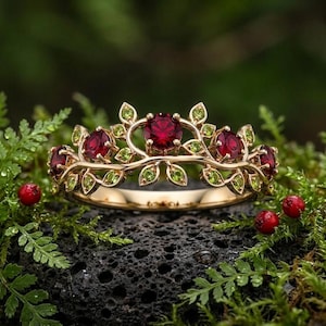 May include: A gold ring featuring a central round red gemstone flanked by two smaller round red gemstones, all set within a band designed with intricate gold leaves and accented with small, round, light green peridot-colored stones.
