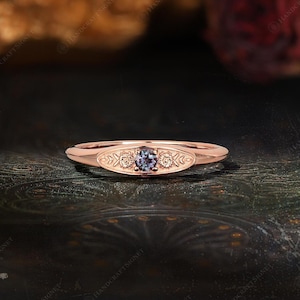May include: A rose gold ring featuring a central round-cut purple-blue gemstone set within an oval-shaped plate. The plate is decorated with engraved leaf and floral patterns on either side of the stone, set on a smooth, polished band.
