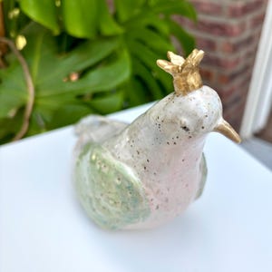Könnte beinhalten: Keramik-Vogelfigur mit goldener Krone, hellgrünem Flügel und rosa Brust. Der Vogel hat einen gesprenkelten, cremefarbenen Körper und einen goldenen Schnabel. Die Figur steht auf einer weißen Oberfläche.