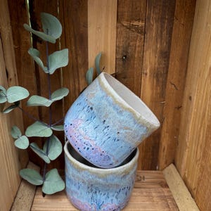 Könnte beinhalten: Ein Set aus drei Keramikbechern mit gesprenkelter, matter Oberfläche. Die Becher sind in Creme-, Hellgrau- und Dunkelgrautönen gehalten. Jeder Becher hat ein schlichtes, minimalistisches Design.