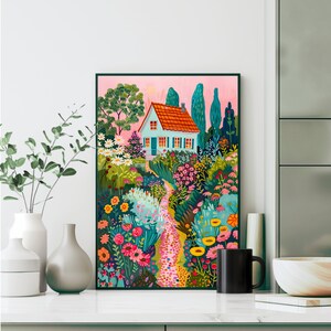 May include: A framed art print showing a garden path leading to a light blue house with an orange roof. The artwork features vibrant flowers, trees, and a pink sky, creating a whimsical scene. The print is displayed on a white surface.