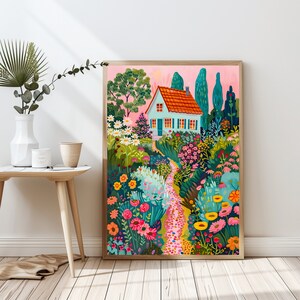 May include: A framed art print showing a garden scene with a light blue house, orange roof, and a winding path. The garden is filled with vibrant flowers and greenery, set against a pink sky. The artwork is displayed on a wooden floor.