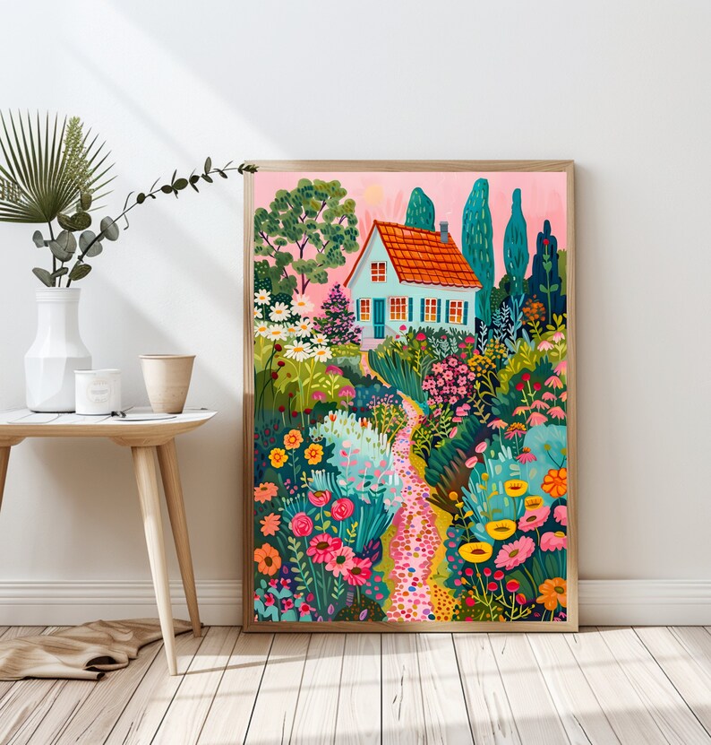 May include: A framed art print showing a garden scene with a light blue house, orange roof, and a winding path. The garden is filled with vibrant flowers and greenery, set against a pink sky. The artwork is displayed on a wooden floor.